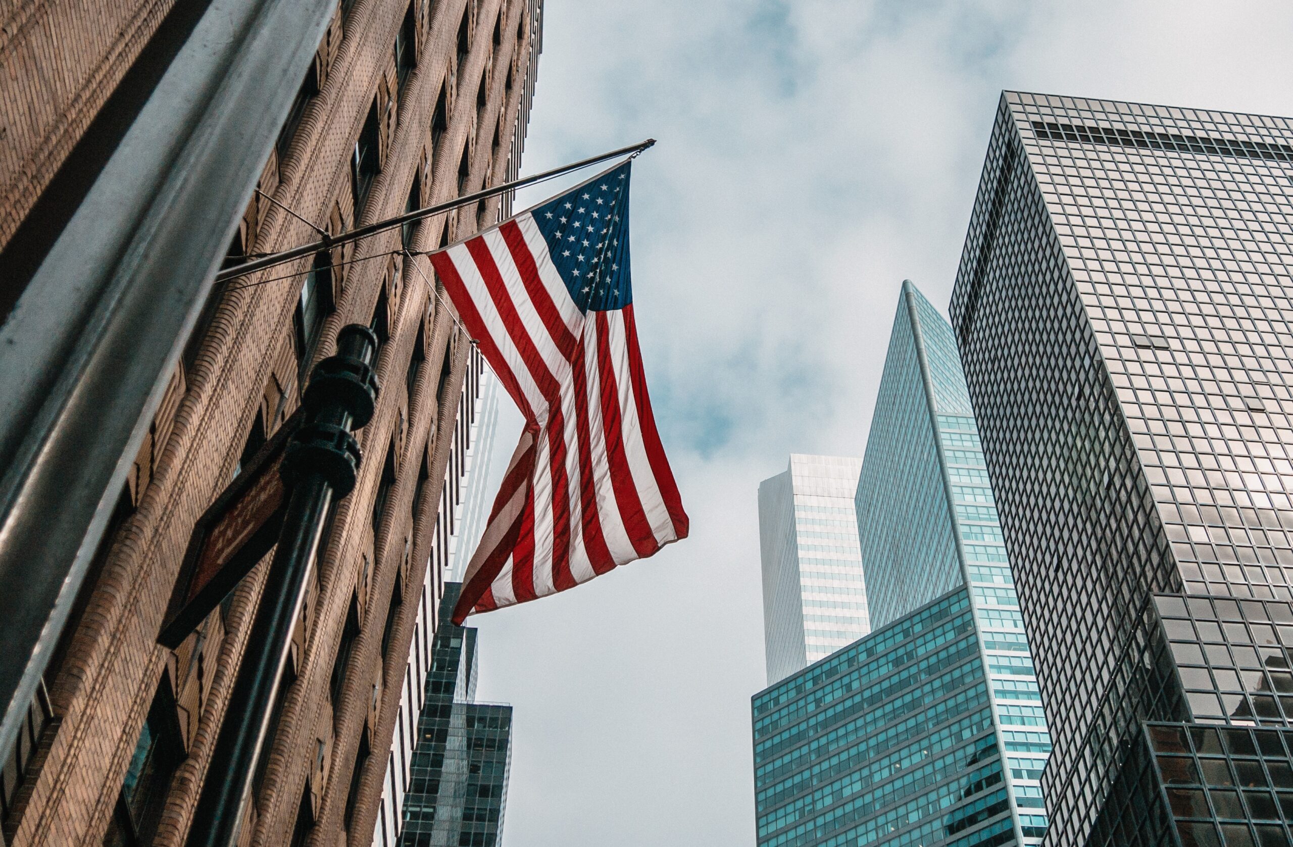 A low angle shot of the USA or United States of America flag on a flagpole near skyscrapers under a cloudy sky