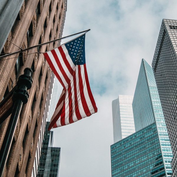 A low angle shot of the USA or United States of America flag on a flagpole near skyscrapers under a cloudy sky