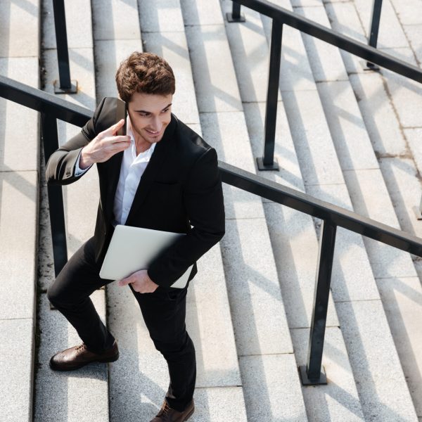 Image of happy young businessman walking outdoors. Talking by phone holding laptop computer. Looking aside.